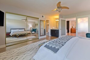 Un dormitorio moderno con un espejo grande, una cama con una colcha estampada, un ventilador de techo y vista a otro cuarto con baño.