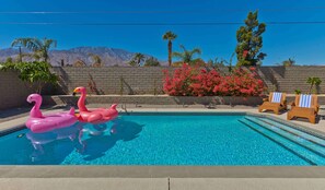 A swimming pool with two pink flamingo floats, a brick wall, and a row of wooden lounge chairs. - Modern Home w Private Pool Spa Pets Welcome (Palm Springs)
