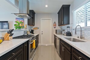 A modern kitchen with dark cabinets, a central island, a stove, and a large window with blinds. - Modern Home w Private Pool Spa Pets Welcome (Palm Springs)
