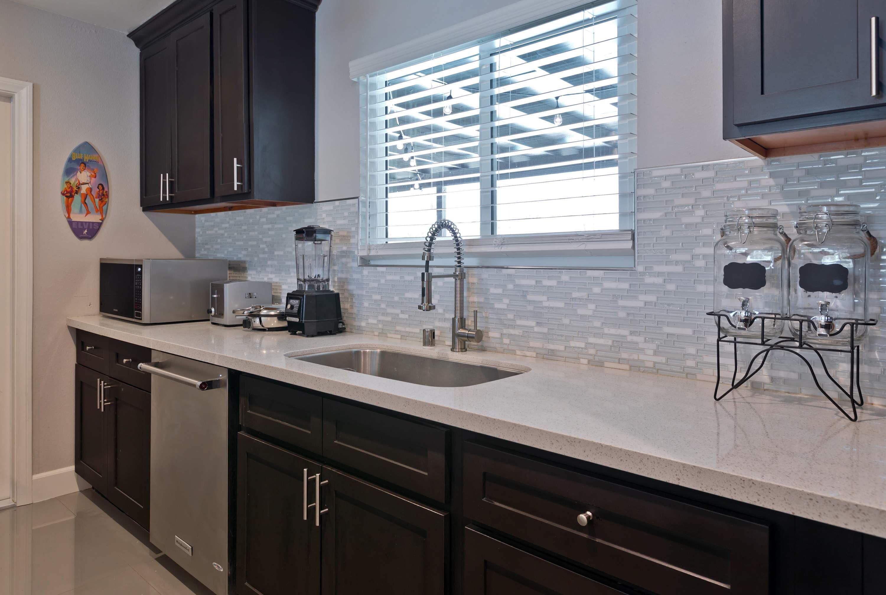 A modern kitchen with dark cabinets, a large window with blinds, a stainless steel sink, and a coffee maker.