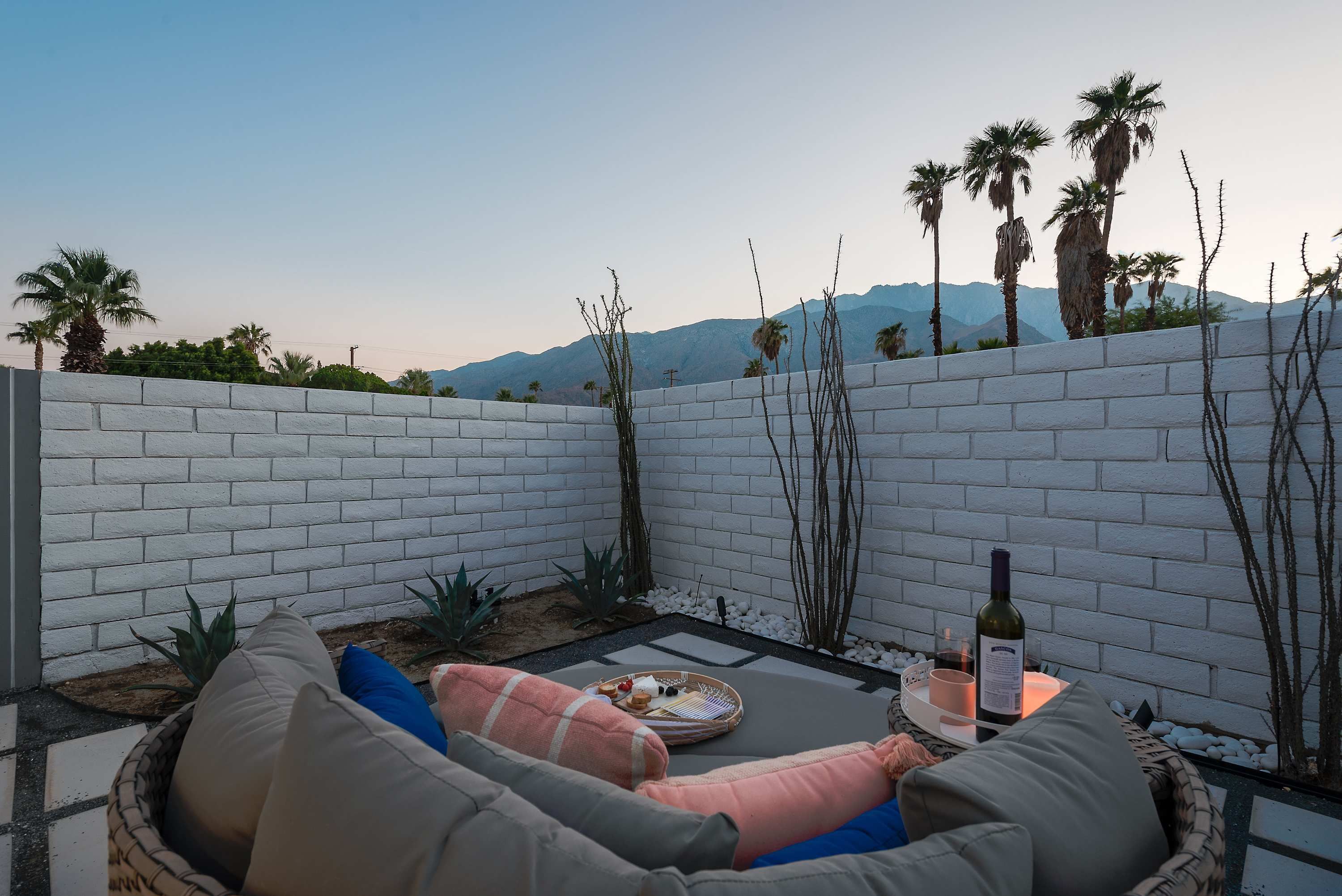 Une terrasse avec un mur en briques blanches, un coin salon avec des coussins, une petite table avec une bouteille et une tasse, et une vue sur des palmiers et des montagnes.