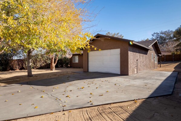 A single-story house with a garage, a driveway with scattered leaves, and a tree with yellow leaves. - Near JT Oasis Hot Tub Cowboy Tub Games (Joshua Tree)