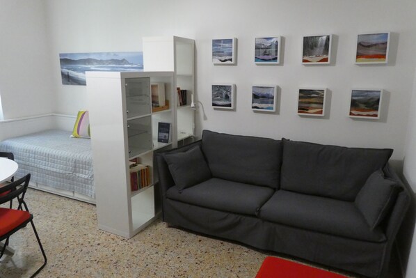 Una habitación con un sofá gris, una mesita con una silla, una estantería y una cama con una manta azul.