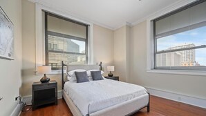 Un dormitorio con una cama, dos mesitas de noche y ventanas grandes con persianas.