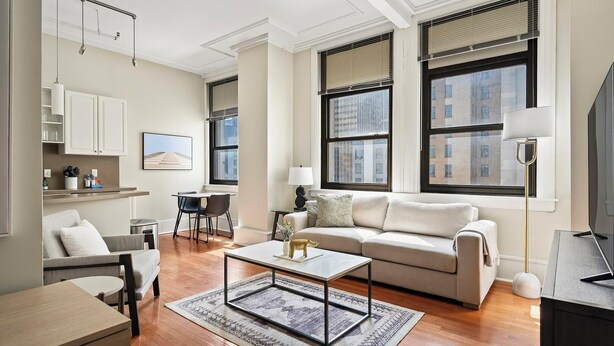 A modern living room with a sofa, coffee table, and dining area. There are large windows with blinds, a TV, and a lamp.