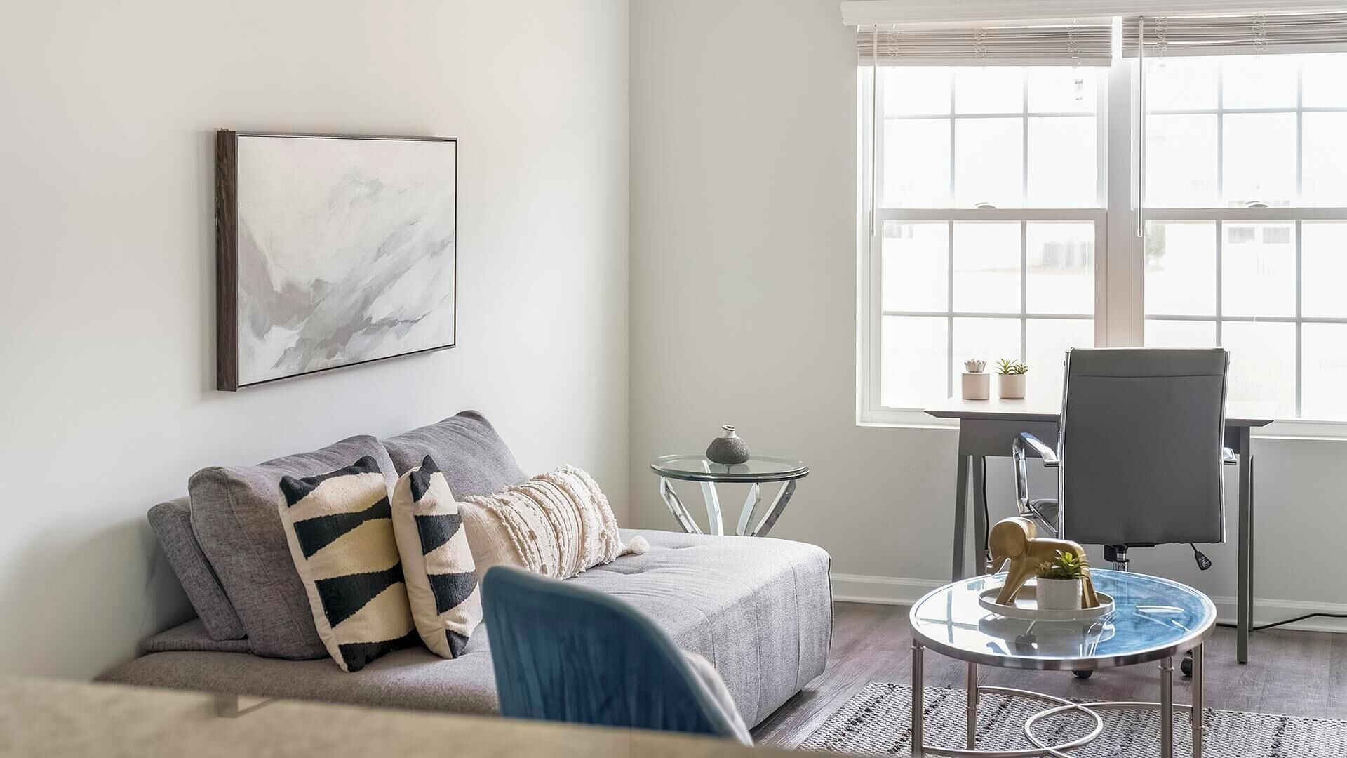 A modern living room with a grey sofa, a glass coffee table, a blue chair, and a window with blinds.