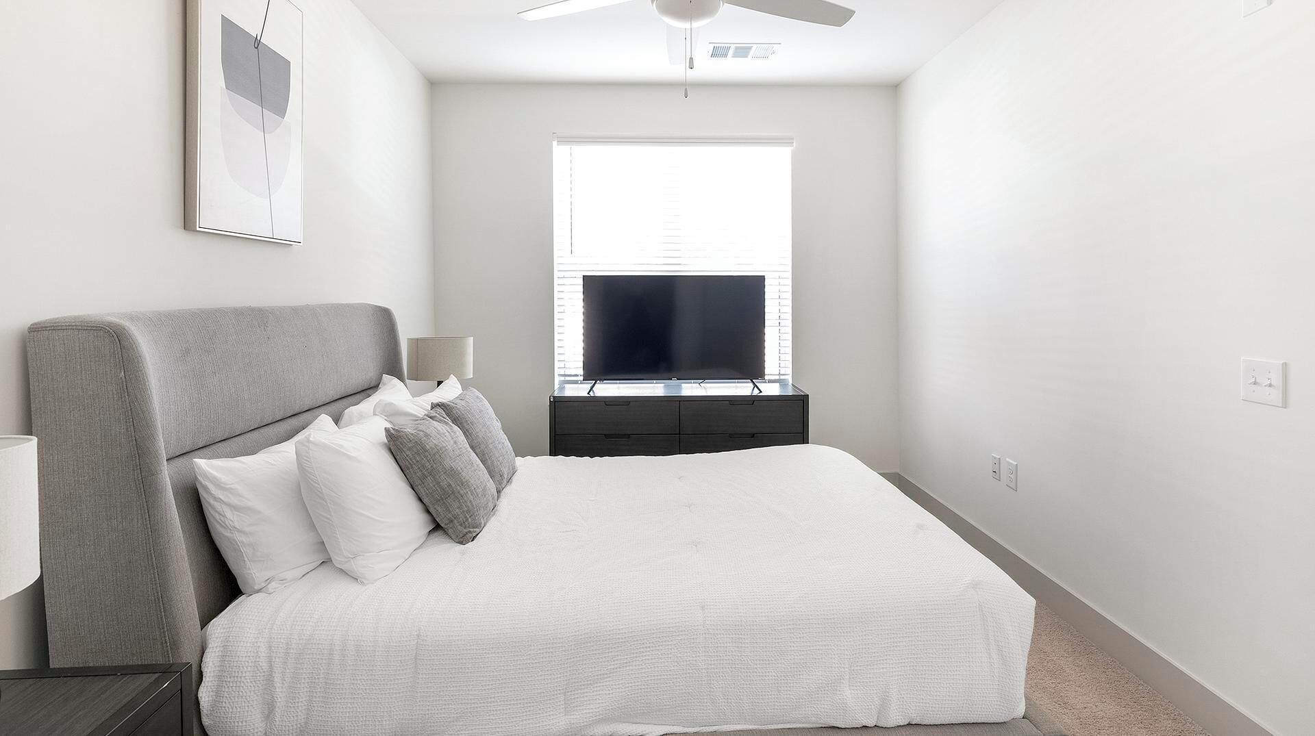 A bedroom with a bed, a TV, a ceiling fan, and a window with blinds.
