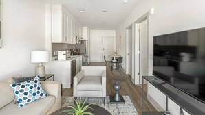 A modern living room with a sofa, coffee table, and TV. There is a kitchen area with cabinets and a dining table. - Landing | Modern 1BD, Gym, Clubhouse (Huntsville)