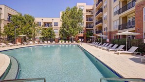 A rectangular pool with clear blue water, surrounded by lounge chairs and umbrellas, with multi-story buildings in the background. - Landing | Modern 1BD, Courtyard, Gym (Cary)