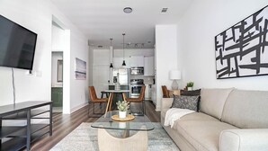 En moderne stue med sovesofa, glasbord, fjernsyn ophængt på væggen og et køkken i baggrunden.