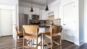 A modern kitchen with white cabinets, a central island, two bar stools, pendant lights, and a stainless steel refrigerator. - Landing | Modern 2BD, Pool, Rooftop Lounge (Cary)