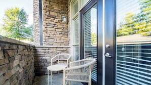 A stone exterior wall, a glass door with a metal handle, and two wicker chairs on a concrete floor. - Landing | Modern 2BD, Pool, Rooftop Lounge (Cary)