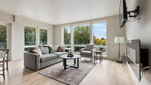 A modern living room with a grey sectional sofa, a round coffee table, and a grey armchair. There are large windows offering a view of trees and a clear sky. - Landing | Amazing 2BD, Gym, Pool (Portland)
