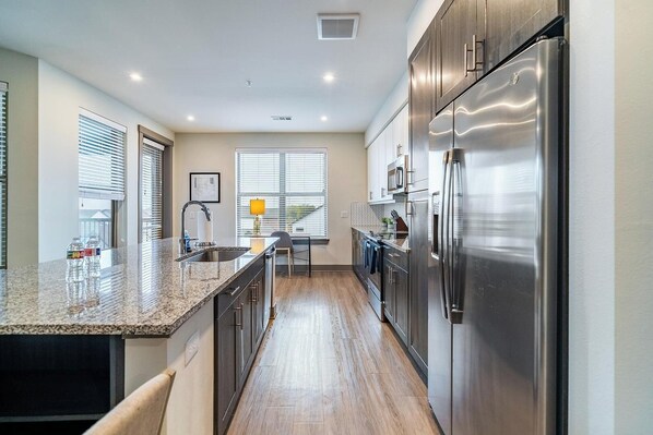 A modern kitchen with stainless steel appliances, a granite countertop, and a view of the outside through large windows.