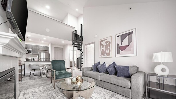 A modern living room with a grey sofa, a green armchair, and a glass coffee table. There is a kitchen in the background with white cabinets and a black staircase. - Landing | Stylish 2BD, Pool, Clubhouse (Cincinnati)