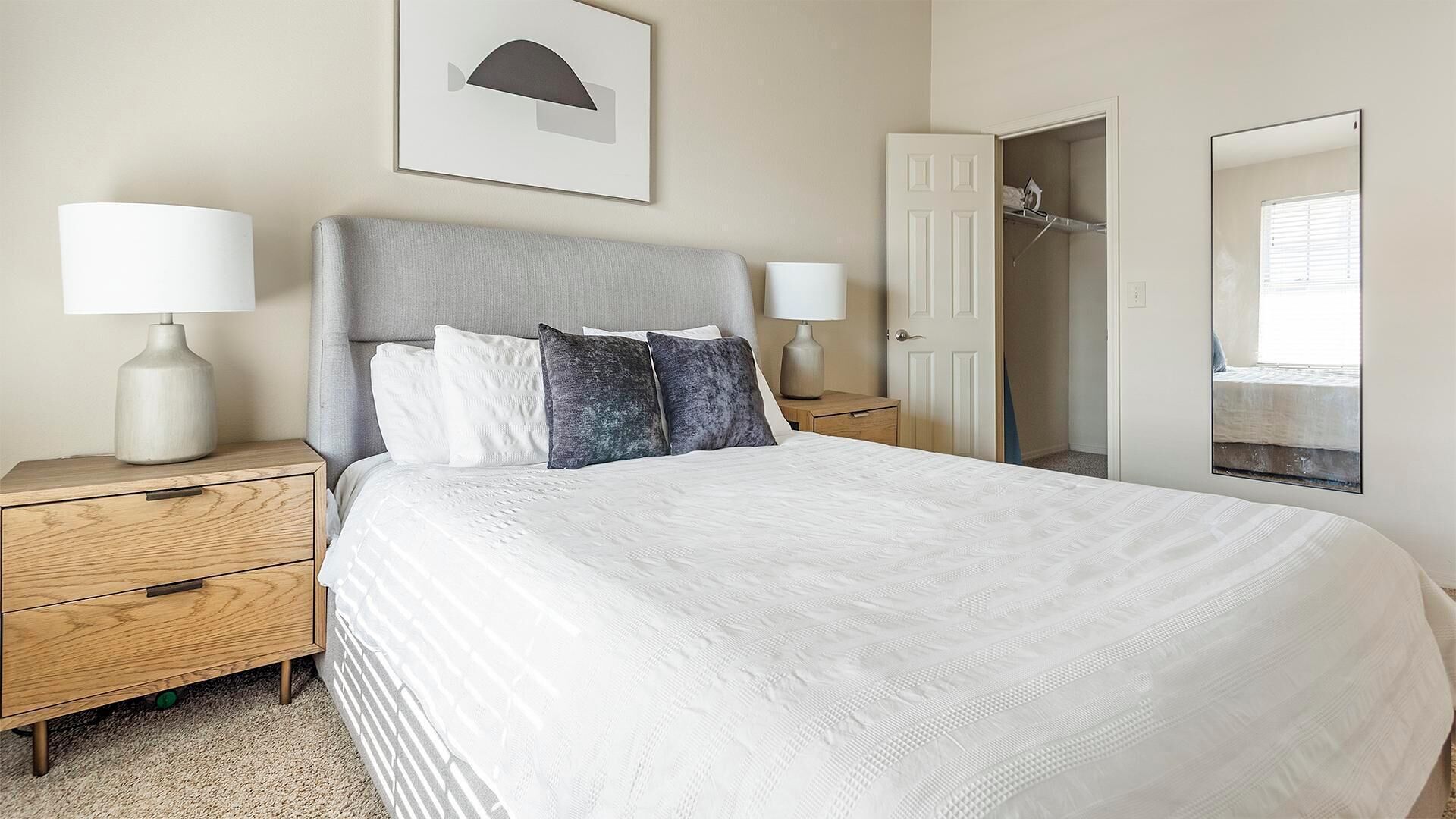 A neatly made bed with a headboard, two bedside tables with lamps, a wooden nightstand, a framed picture on the wall, and a closet with a mirror.