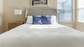A neatly made bed with a headboard, a bedside table with a lamp, and a window with blinds.