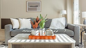 Ein Wohnzimmer mit einem grauen Sofa, weißen Kissen, einem Couchtisch mit einer Pflanzentafel und einer Lampe.