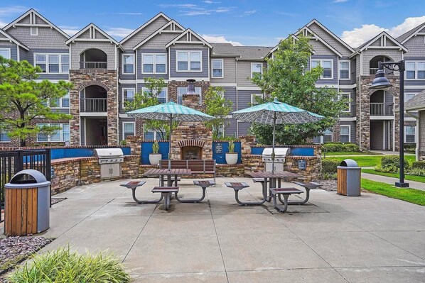 A patio area with picnic tables and barbecue grills, surrounded by stone walls and a row of townhouses.