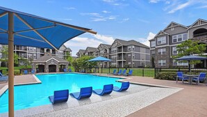 A swimming pool with blue chairs and umbrellas, surrounded by multi-story buildings with balconies. - Landing | Amazing 2BD, Courtyard, Gym (Broken Arrow)