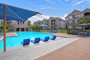 Una piscina con sillas y sombrillas azules, rodeada de edificios de varios pisos con balcones.