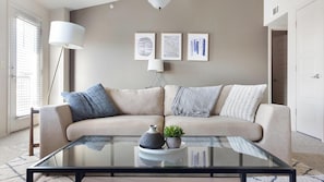 A living room with a beige sofa, a glass coffee table, and a floor lamp. There are three framed artworks on the wall. - Landing | Modern 2BD, Pool, Courtyard (Jenks)