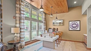 A modern hotel room with a wooden ceiling, a dining area with a white table, and a large window with blinds. - Landing | Modern 2BD, Pool, Gym (Jenks)