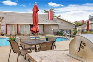 A poolside area with a table and chairs, a red umbrella, and a barbecue grill. - Landing | Beautiful 1BD, Gym, Courtyard (Grand Prairie)