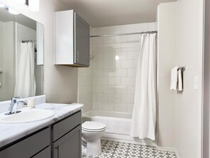 Un baño moderno con un lavabo, espejo y retrete blancos, una ducha con cortina y piso de baldosas.