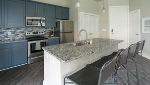A modern kitchen with dark blue cabinets, stainless steel appliances, a granite countertop, and a bar area with stools. - Landing | Stunning 1BD, Gym, Pool (Houston)
