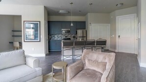 A modern living room with a sofa and armchair, a kitchen in the background, and a dining area with chairs. - Landing | Stunning 1BD, Gym, Pool (Houston)