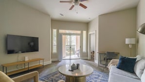A spacious living room with a flat-screen TV, a ceiling fan, a round coffee table, and a large window with a view of the outdoors.