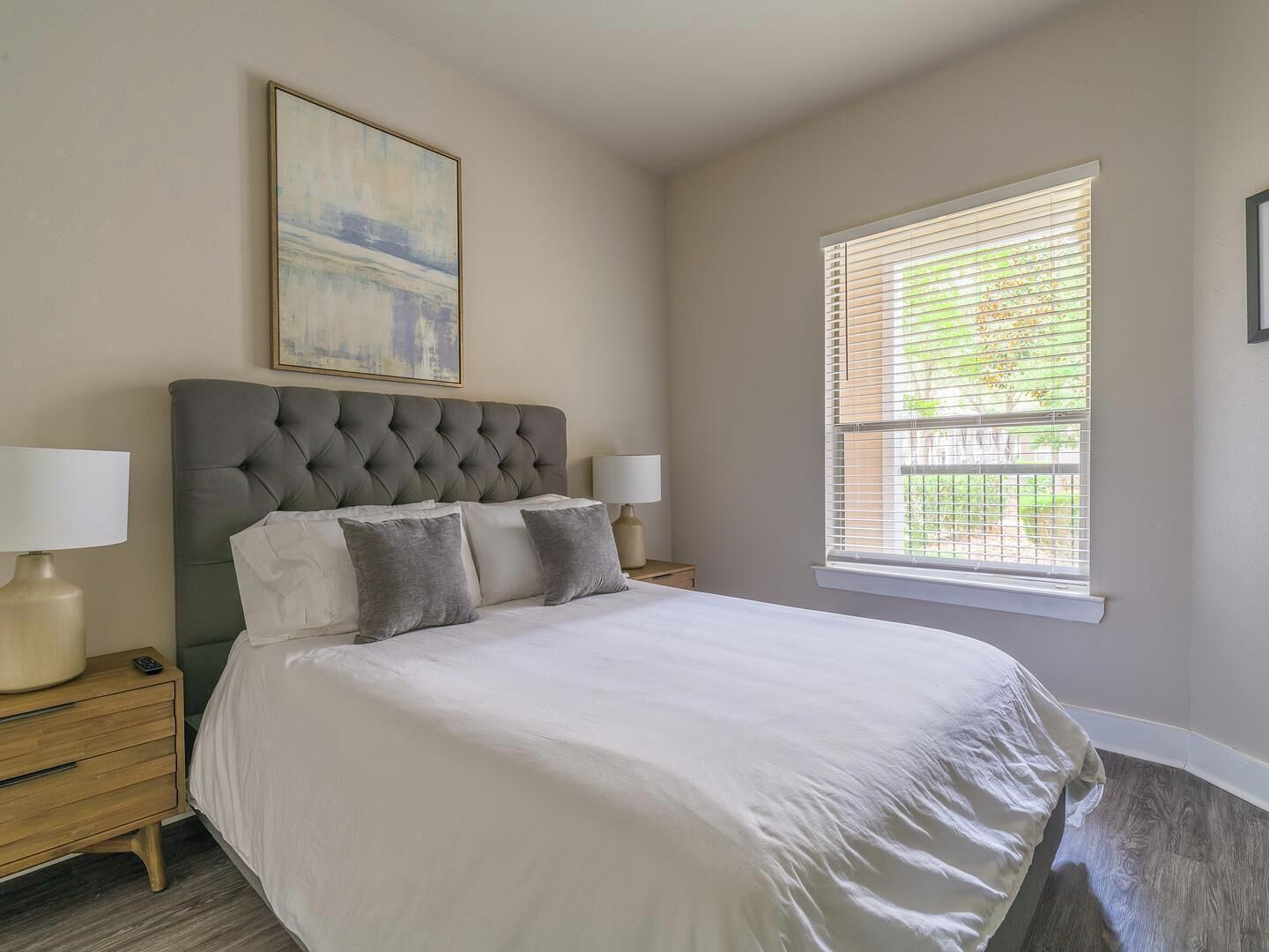 A bedroom with a tufted headboard, a large bed with white linens, two bedside lamps, a nightstand, a window with blinds, and a framed abstract painting on the wall.