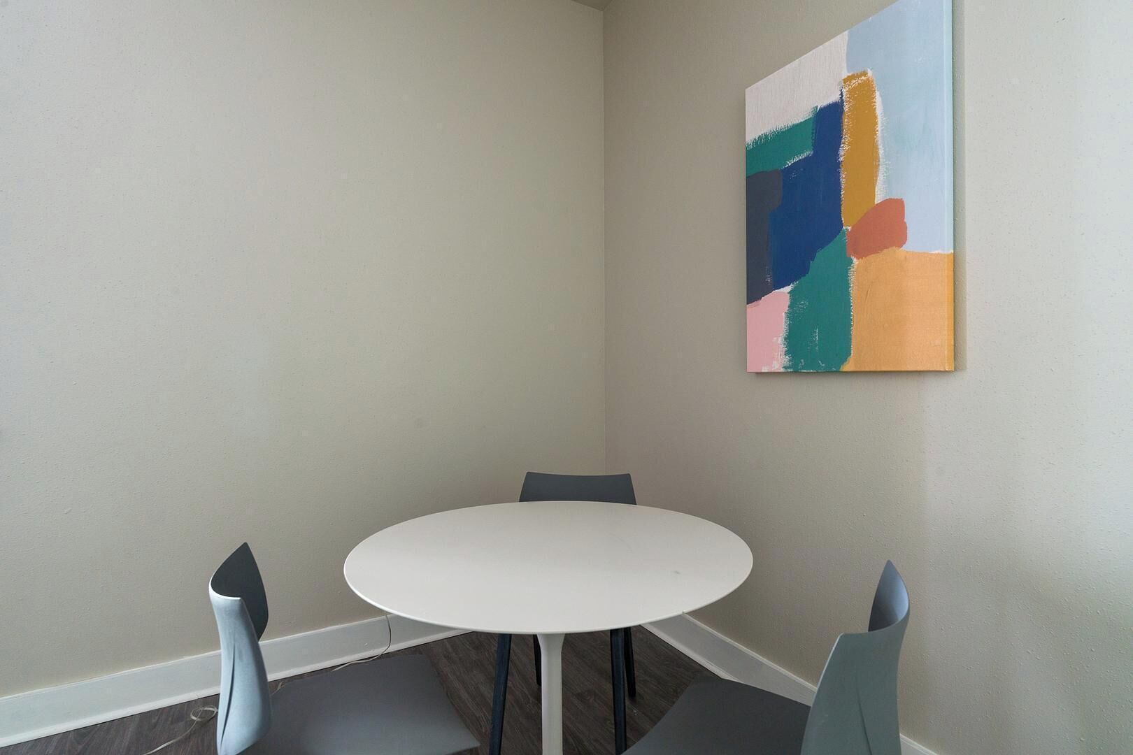Een kleine, ronde tafel met vier stoelen in een hoek van een kamer met kale muren en een kleurrijk abstract schilderij.