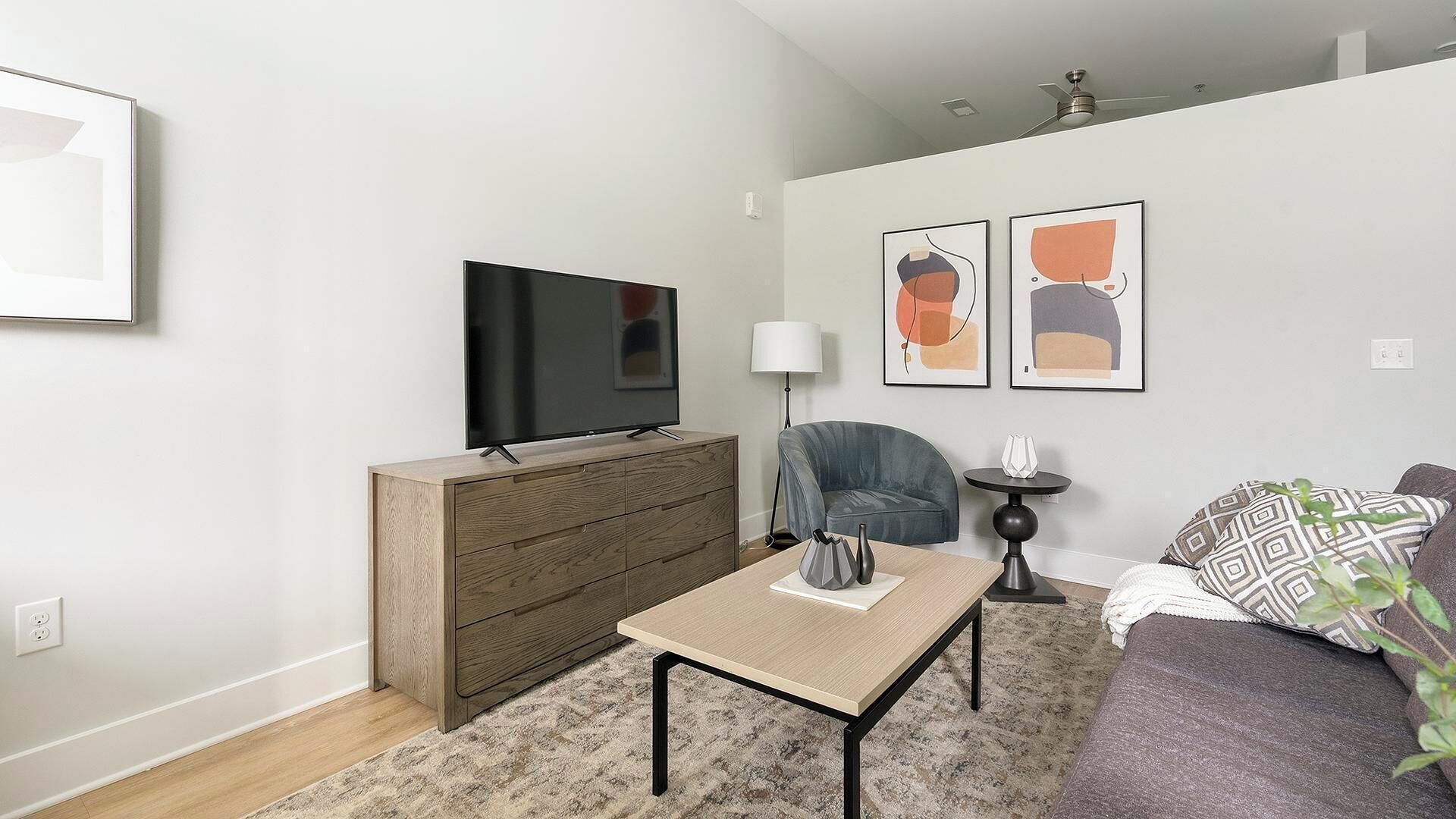 A modern living room with a flat-screen TV on a wooden cabinet, a grey sofa, a coffee table, and abstract art on the wall.