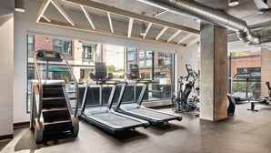 A modern gym with treadmills, a stair climber, and exercise bikes, large windows, and a concrete ceiling.