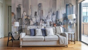 Un salon avec un canapé gris, deux tables d’appoint et une fresque murale représentant un paysage urbain.