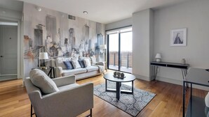 Un salon moderne avec un canapé, un fauteuil et une table basse, une fresque représentant un paysage urbain sur le mur, et un balcon avec vue.