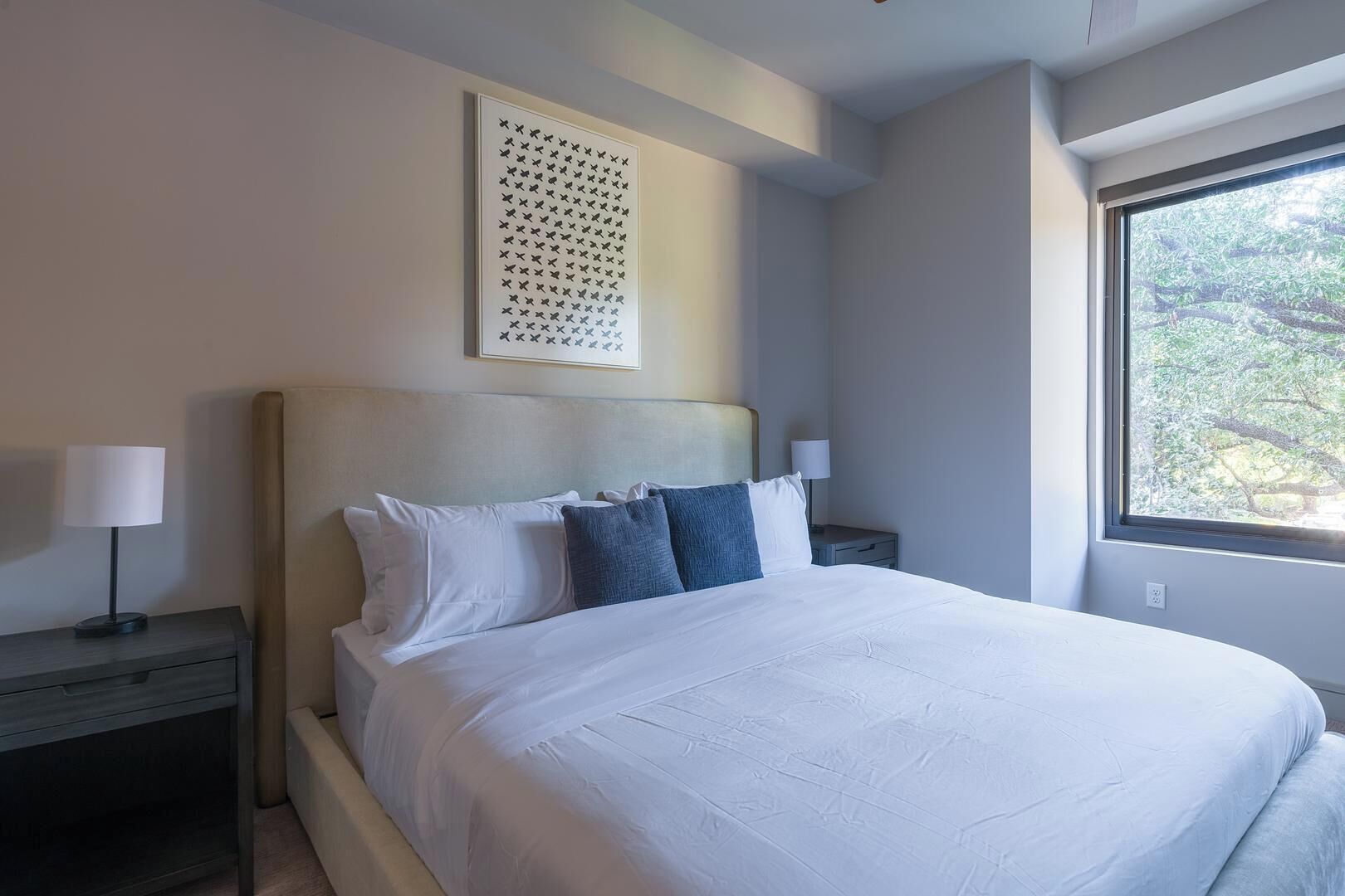 A bedroom with a large bed, two bedside tables, a wall-mounted artwork, and a window with a view of trees.