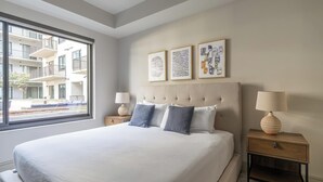 Une chambre d’hôtel moderne dotée d’un grand lit, de deux tables de chevet et d’une fenêtre donnant sur la façade de l’immeuble.