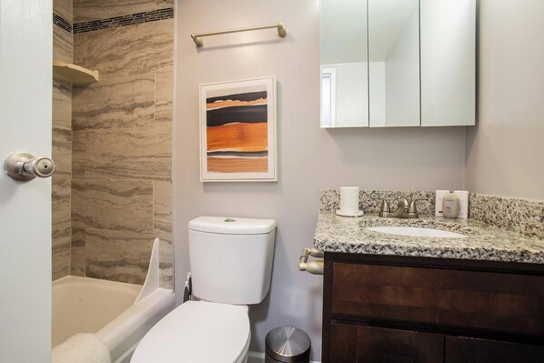 A bathroom with a marble wall, a toilet, a sink with a granite countertop, and a mirror above the sink. - Landing | Beautiful Studio, Gym, Game Room (Fort Wayne)
