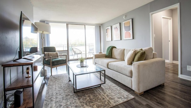 A modern living room with a sofa, coffee table, and a chair. There is a large window providing natural light and a view of the outdoors. - Landing | Beautiful Studio, Gym, Game Room (Fort Wayne)