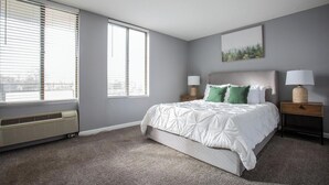 A bedroom with a bed, two bedside tables with lamps, a window with blinds, and a wall-mounted picture. - Landing | Beautiful Studio, Gym, Game Room (Fort Wayne)