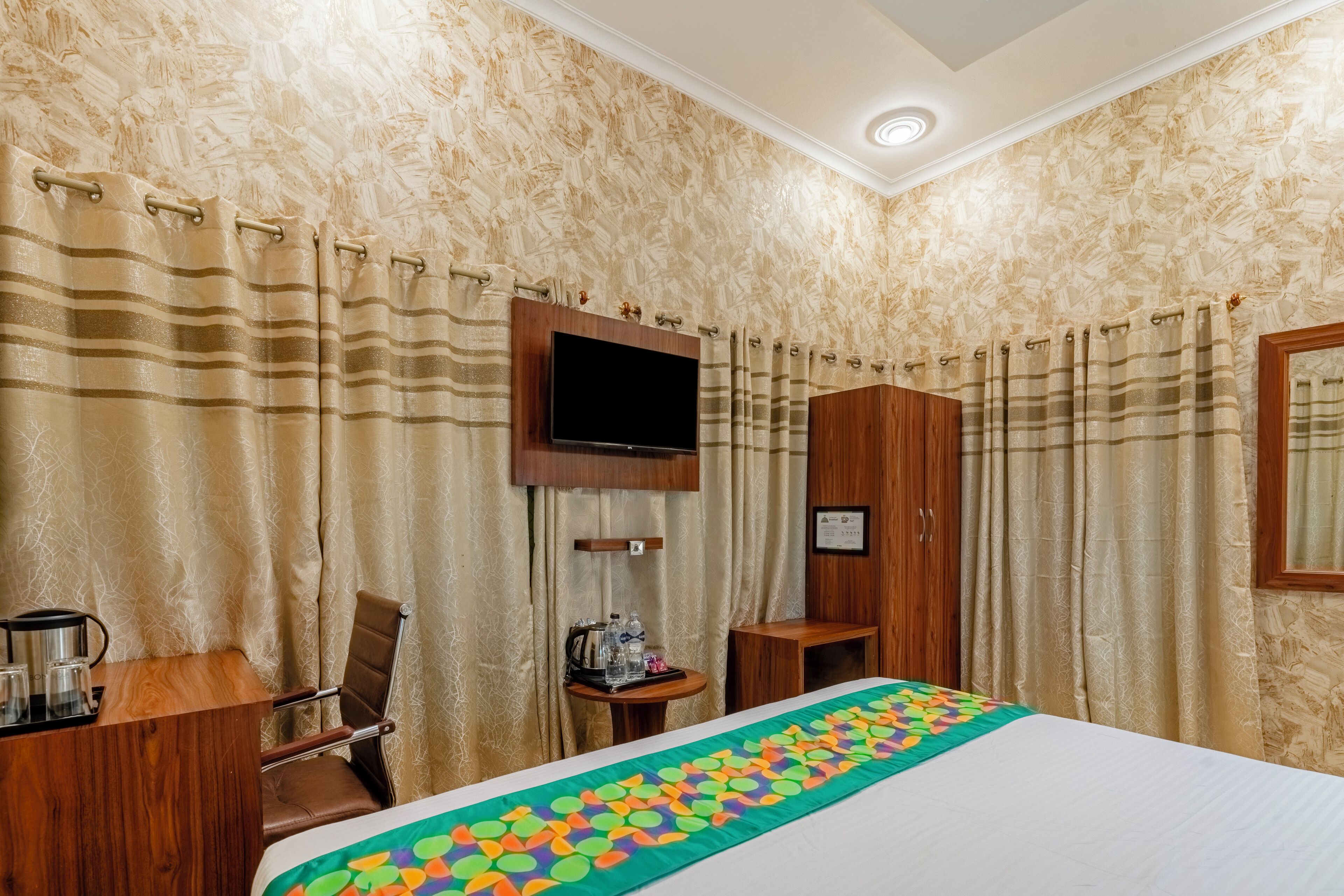 A hotel room with a bed, a TV mounted on the wall, a wooden desk, a chair, and a small table with a kettle and bottles.