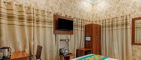 A hotel room with a bed, a TV mounted on the wall, a wooden desk, a chair, and a small table with a kettle and bottles.