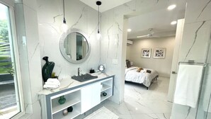 A modern bathroom with a marble vanity, a round mirror, and a bed in the adjacent room.
