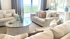 A spacious living room with a large window offering a view of the outdoors, a round glass coffee table, and a sofa set.