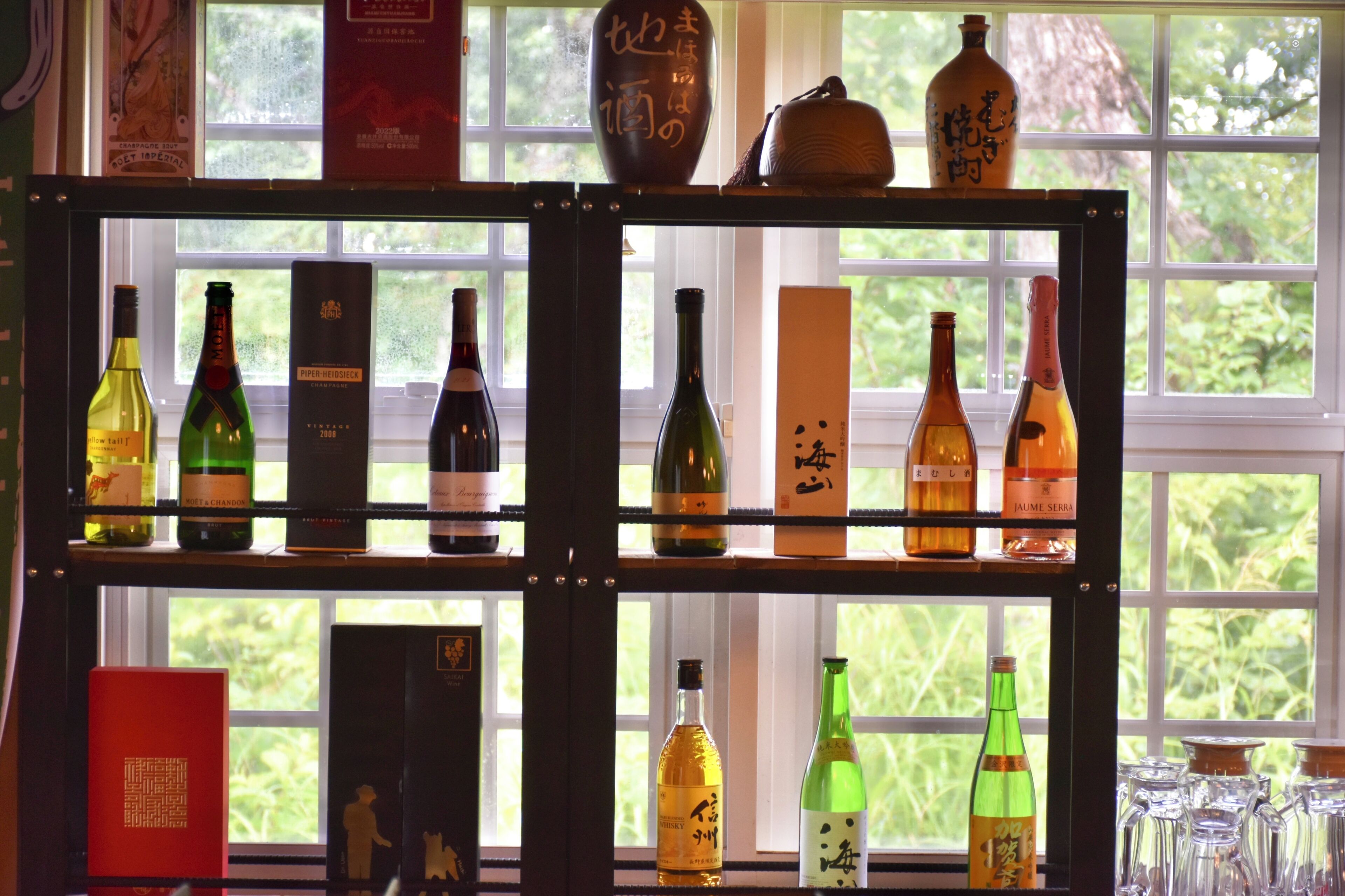 A wine rack with various bottles and boxes, positioned in front of a window with a view of greenery.