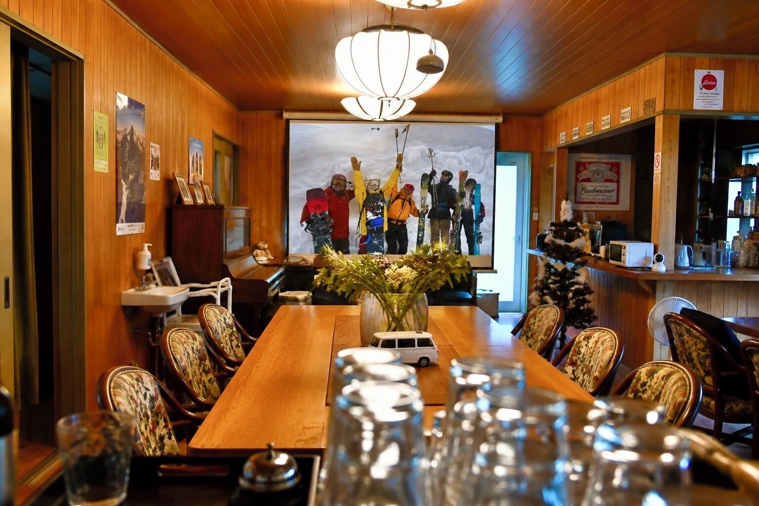 A wooden-paneled room with a long table, chairs, a chandelier, a bar area, and a large poster of skiers.
