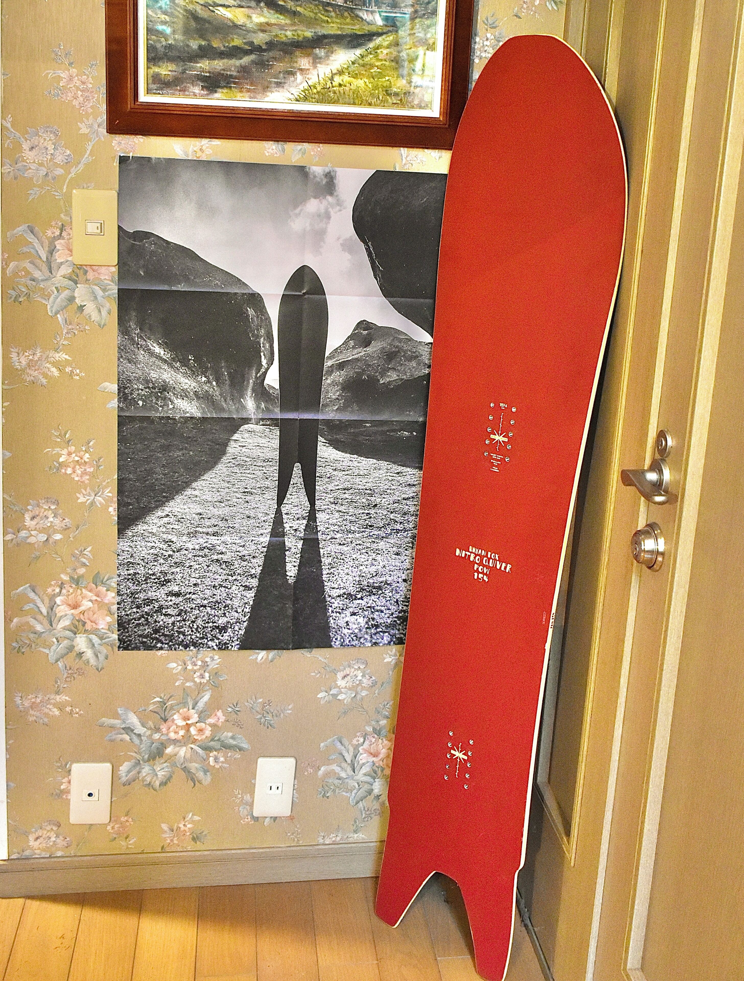 Ein Zimmer mit einem gerahmten Landschaftsbild, einem an der Wand stehenden roten Snowboard und einer Tür mit Knauf.